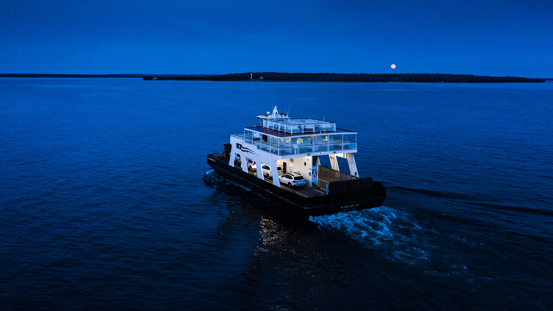 A ferry carrying cars travels across water in the evening with a moonrise visible over trees on a shoreline in the distance.