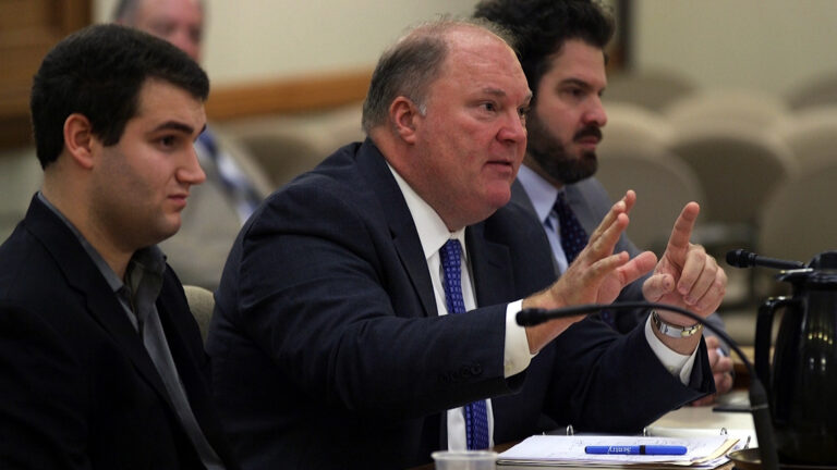 Michael Gableman is seated at a table and speaks into a microphone while holding his hands up, with two men seated on either side of him.