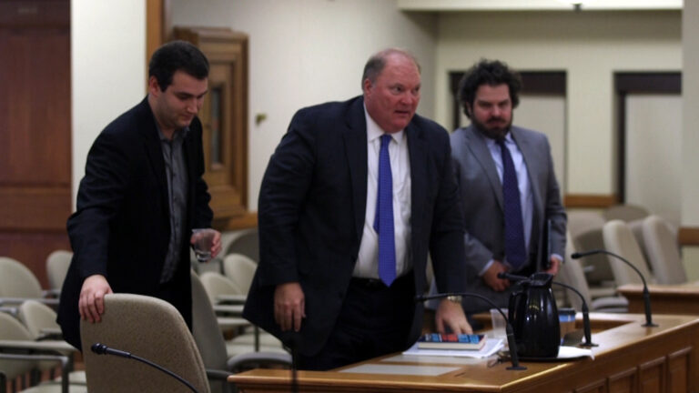 Michael Gableman stands up from a table with two men standing on either side of him.