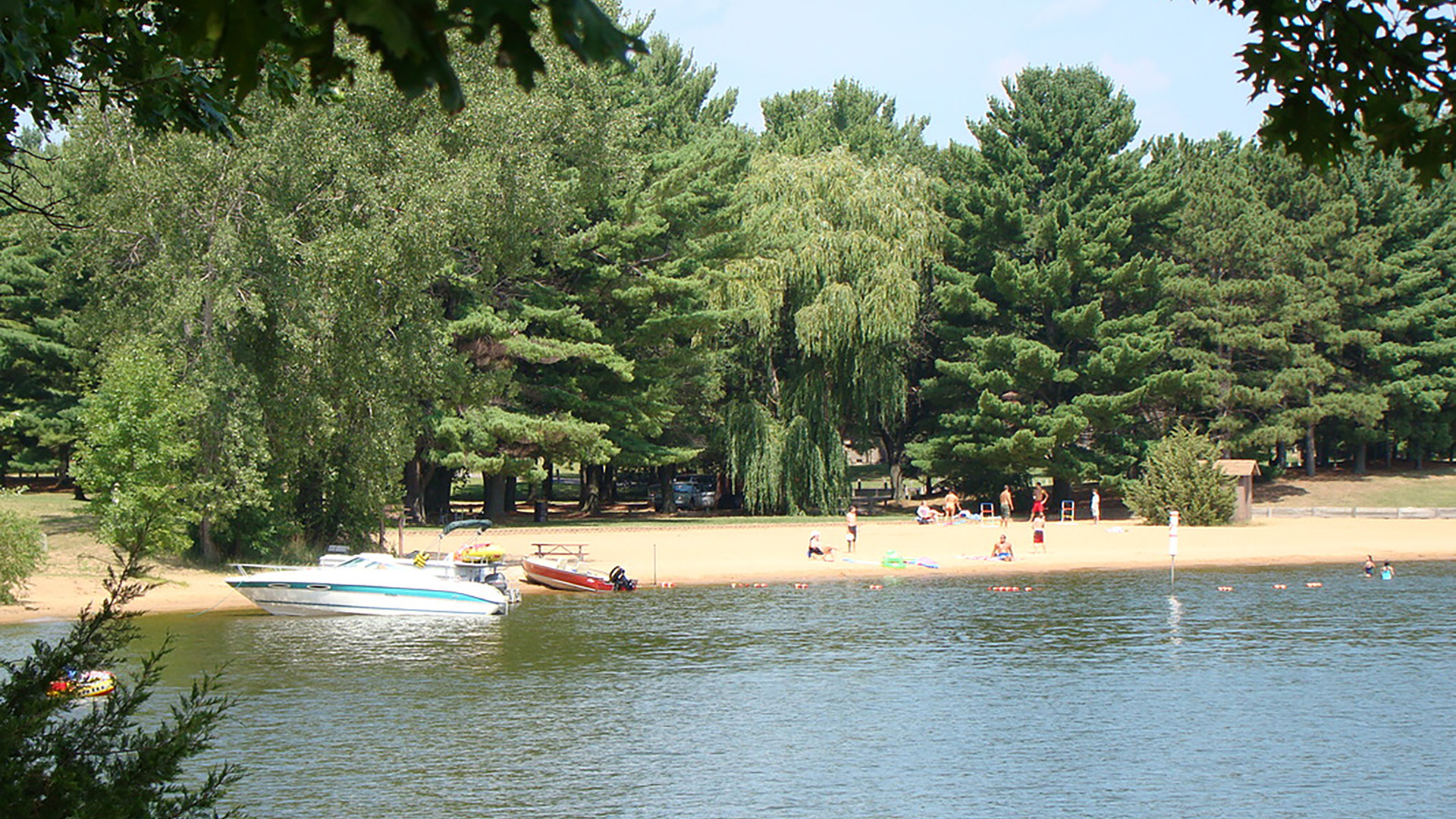 People gather on a sand beach with boats along the shore and trees in the background.