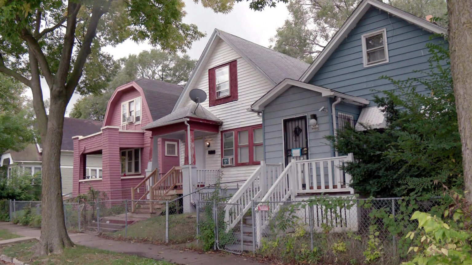 Houses painted red, white and blue stand in a row along a sidewalk with trees in the terrace next to the street and in the background.