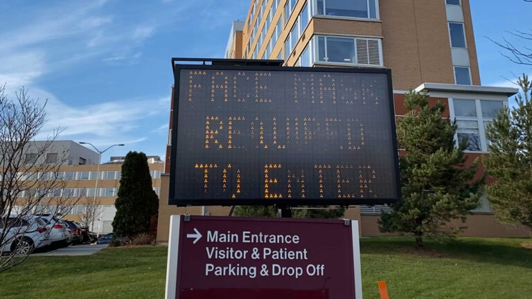 A highway sign reads FACE MASK REQUIRED TO ENTER in front of an entrance sign to a hospital.