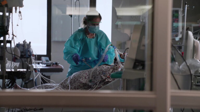 A health care worker attends to an intubated COVID-19 patient in an ICU room as seen through it's glass door.