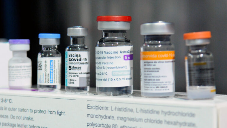Six vials of different COVID-19 vaccines sit in a row on top of a display listing preparation requirements and ingredients.