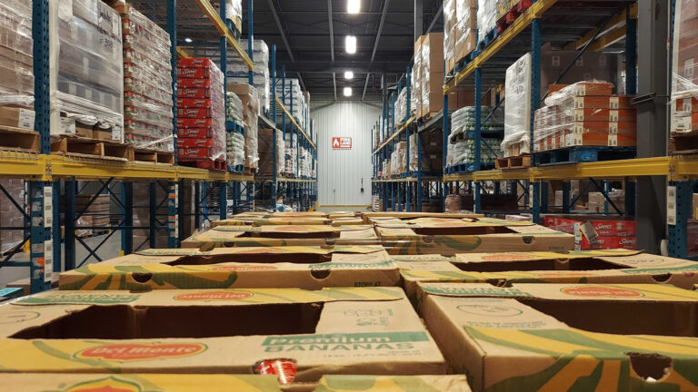 Boxes of food sit stacked in the middle of an aisle in a warehouse with tall shelves filled with boxes on both sides.