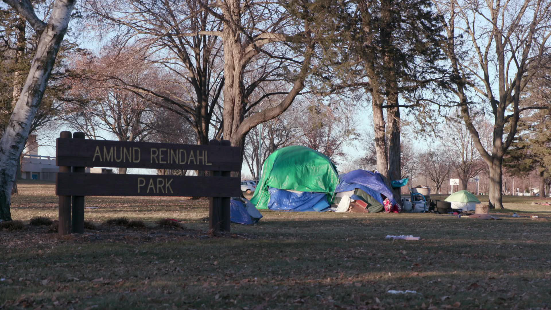 Several tents and surrounding baggage stand under trees and behind a sign reading "Amund Reindahl Park."