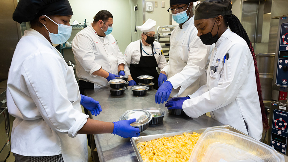 Culinary students stand around a stainless steel table and prepare individual packages of food.