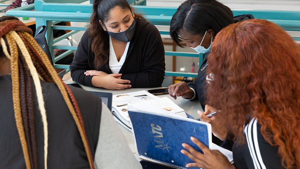 Four students wearing masks sit at a table and look at textbooks and write in notebooks.