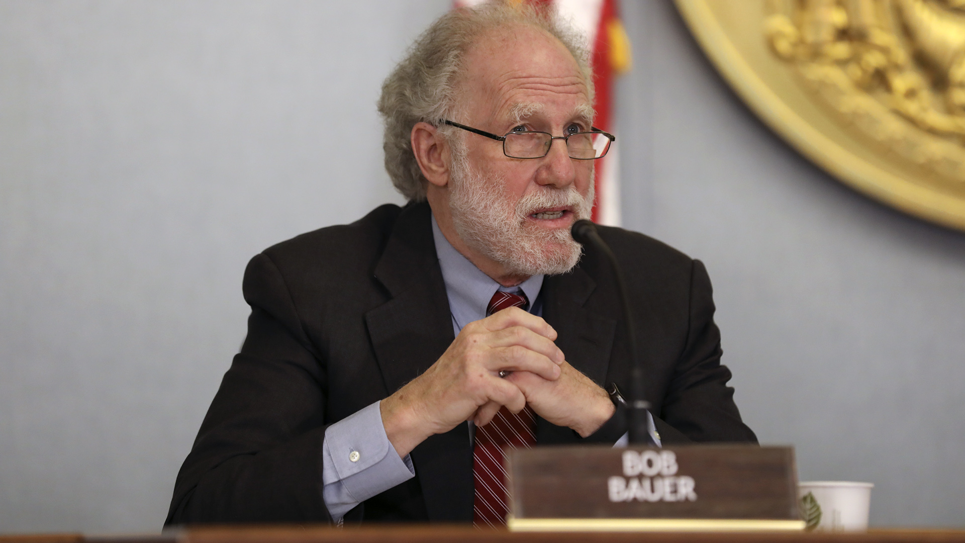 Bob Bauer sits at a conference table facing a microphone and behind a nameplate reading "Bob Bauer."