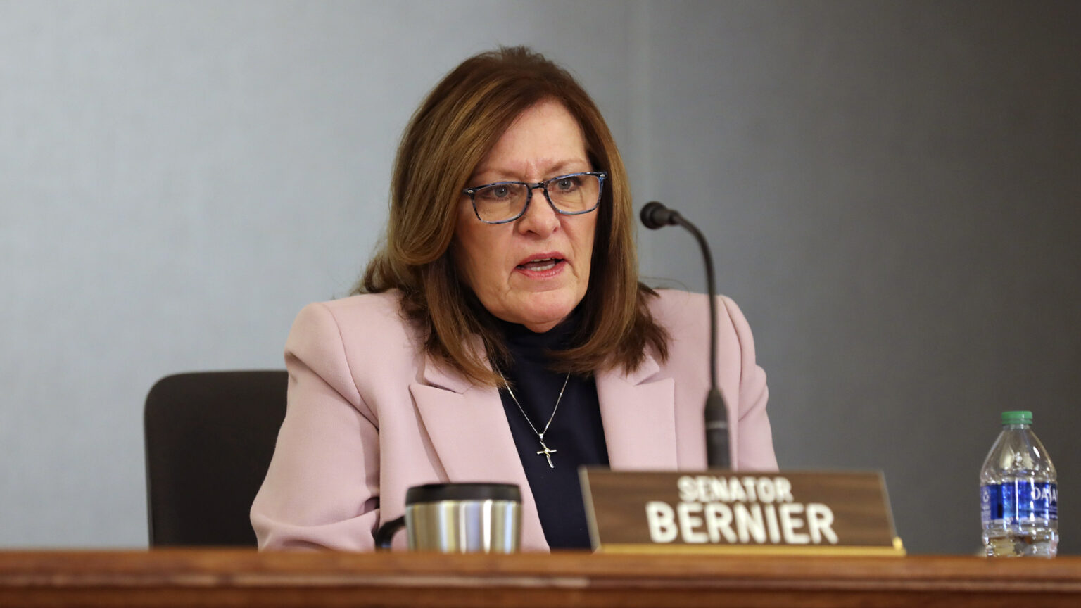Kathleen Bernier sits at a conference table, speaking into a microphone and behind a nameplate reading Senator Bernier.