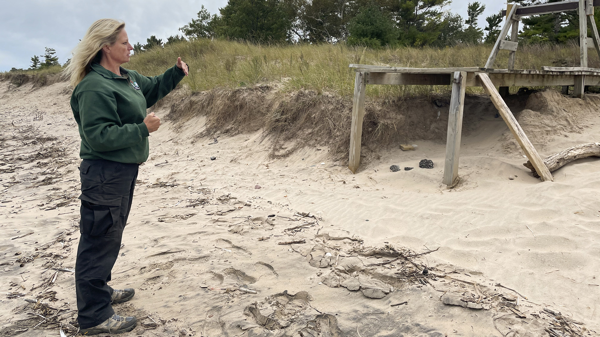 Erin Dembski-Rodriguez stands on a sandy beach and points toward a broken wooden platform.