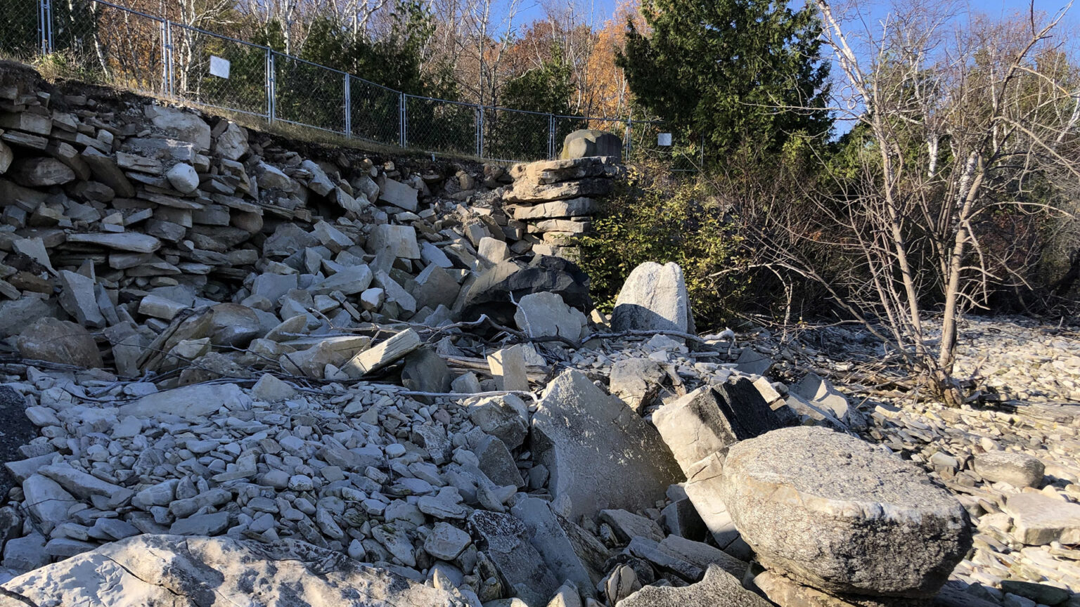 A pile of stones marks where a loose masonry retaining wall is damaged along a shoreline, with trees in the background.