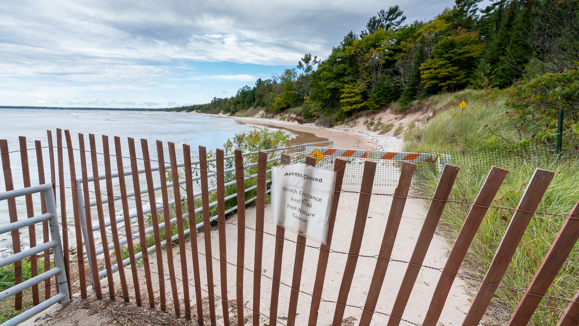 A snow fence with a sign reading "Access Closed, Beach Entrance, Red Trail, Past Nature Center" in front of a sandy slope down to a lakeshore with eroded sandy dunes covered with grass and trees farther inland.