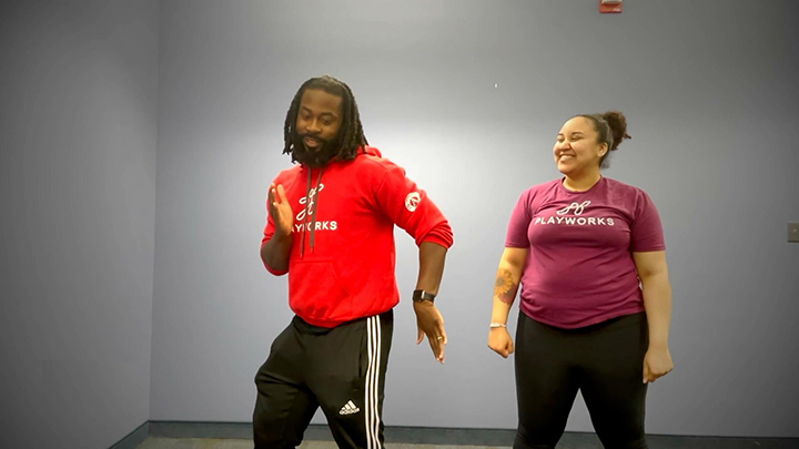 Two people wearing workout gear move together, with one moving and the other looking on and smiling