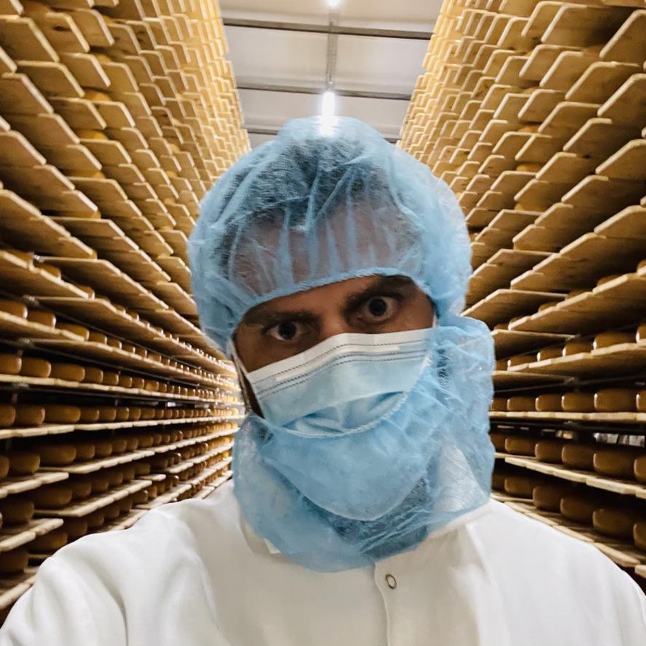 A masked man wearing a hair cap takes a selfie. Shelves of cheese sit behind him. 