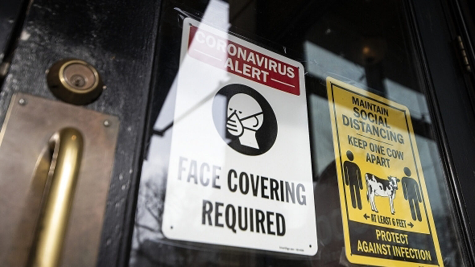 Signs on a business's doorway instruct patrons that face coverings are required and to maintain social distance of 6 feet (or one cow length) apart.