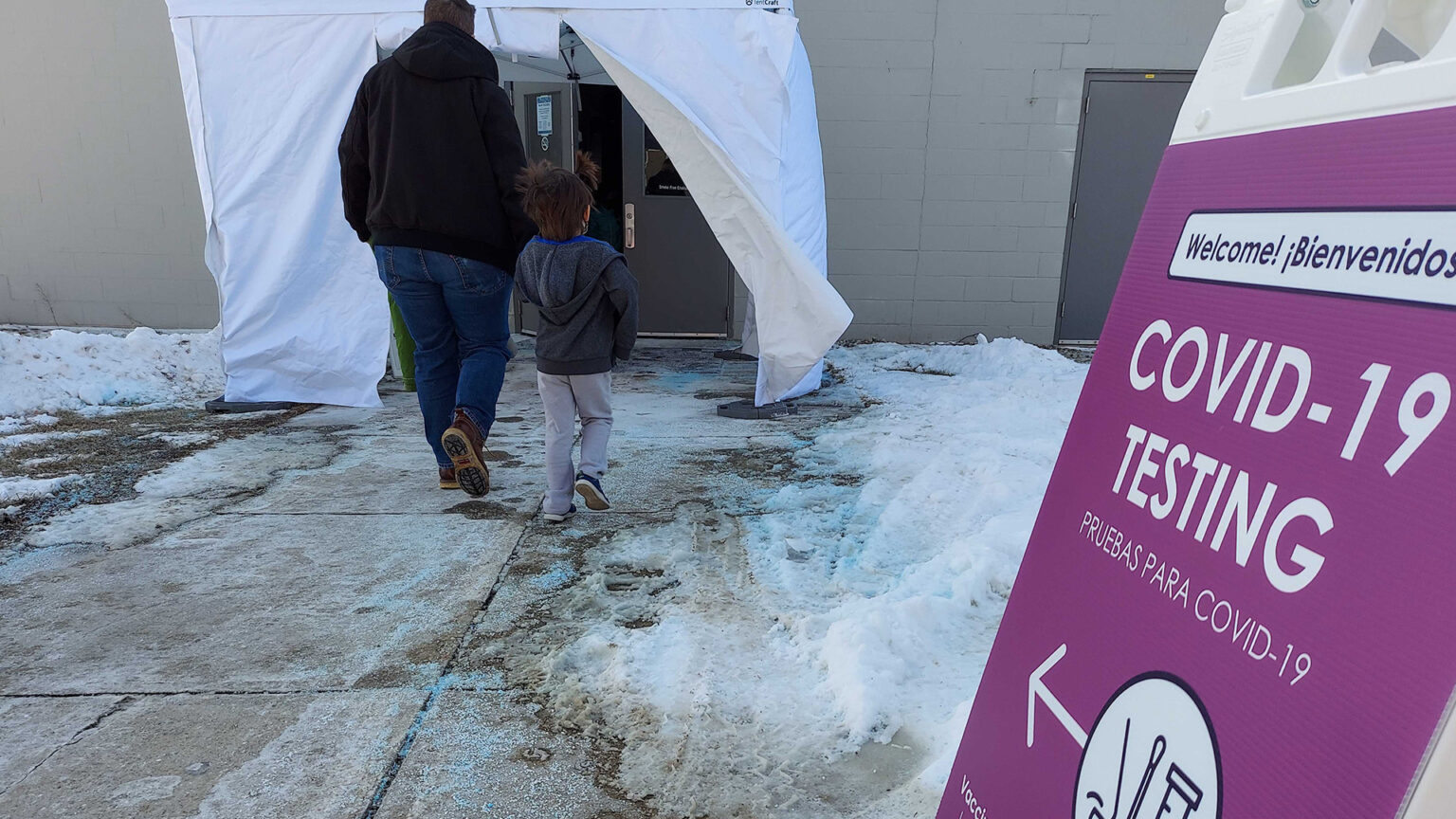 An adult and child walk on an icy sidewalk toward doors covered by an entrance tent, with a sign at right noting the location of COVID-19 testing.