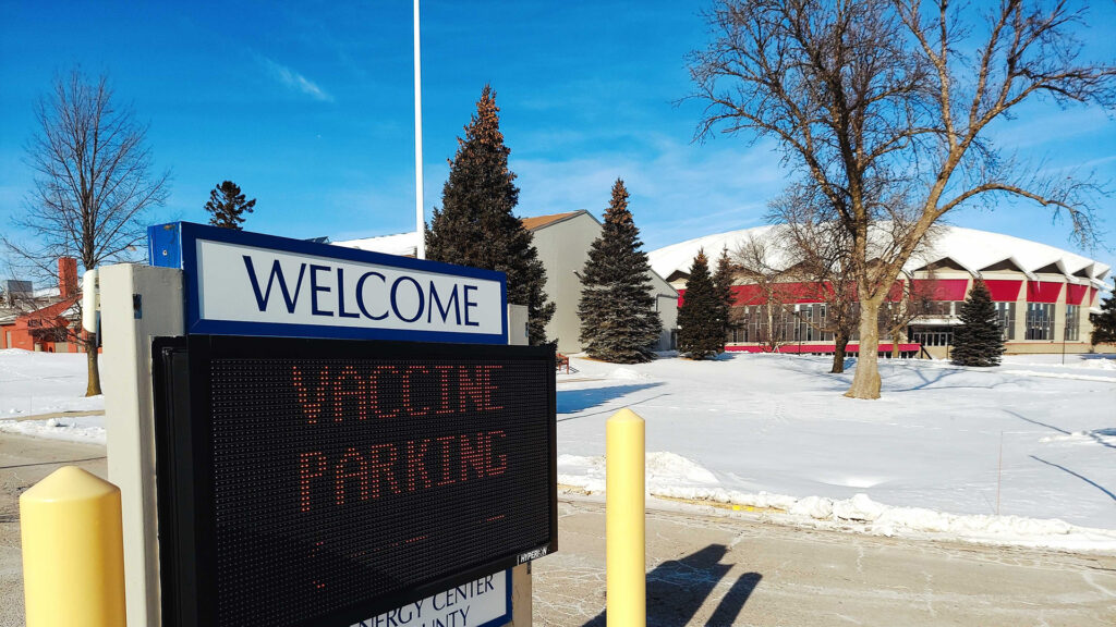 A sign reads "Vaccine Parking," with a coliseum, other buildings, snow-covered ground and trees in the background.