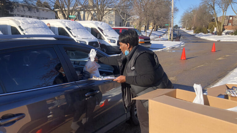 A woman passes a bag through the open window of a stopped car on a road, with snow covered vehicles and lawns in the background.