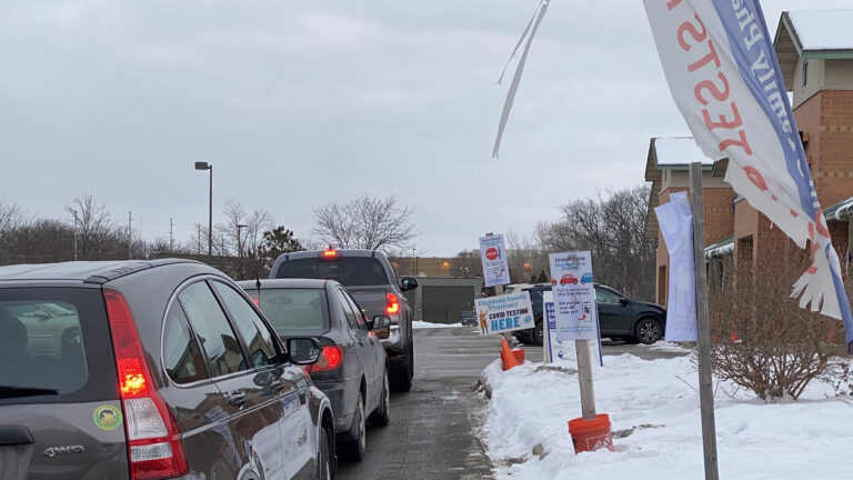 A line of vehicles waits at a COVID-19 testing location next to low office buildings, with signs on a snow-covered lawn providing instructions to drivers.