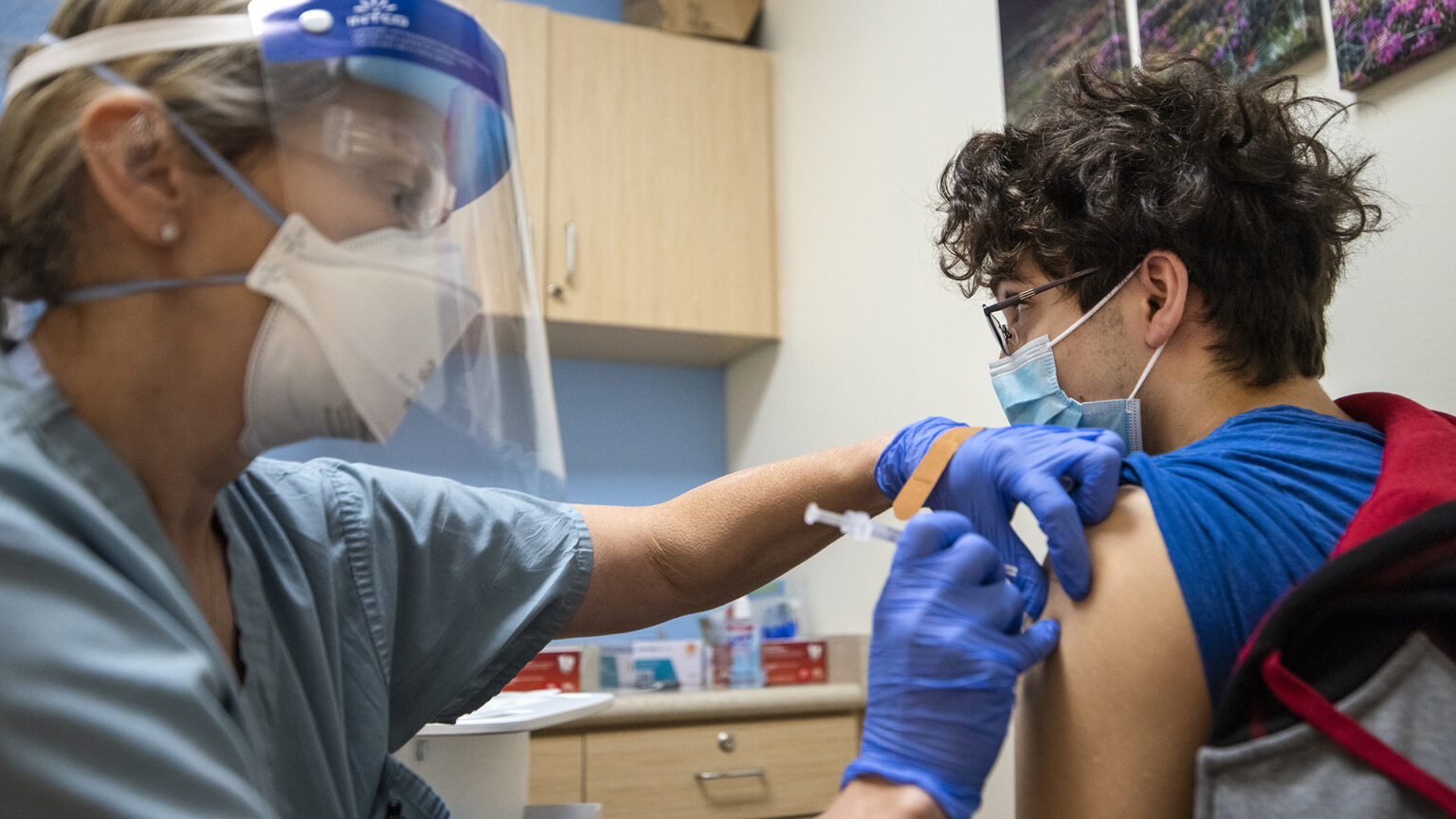 A vaccinator gives a COVID-19 booster shot to a teenager in a medical exam room.