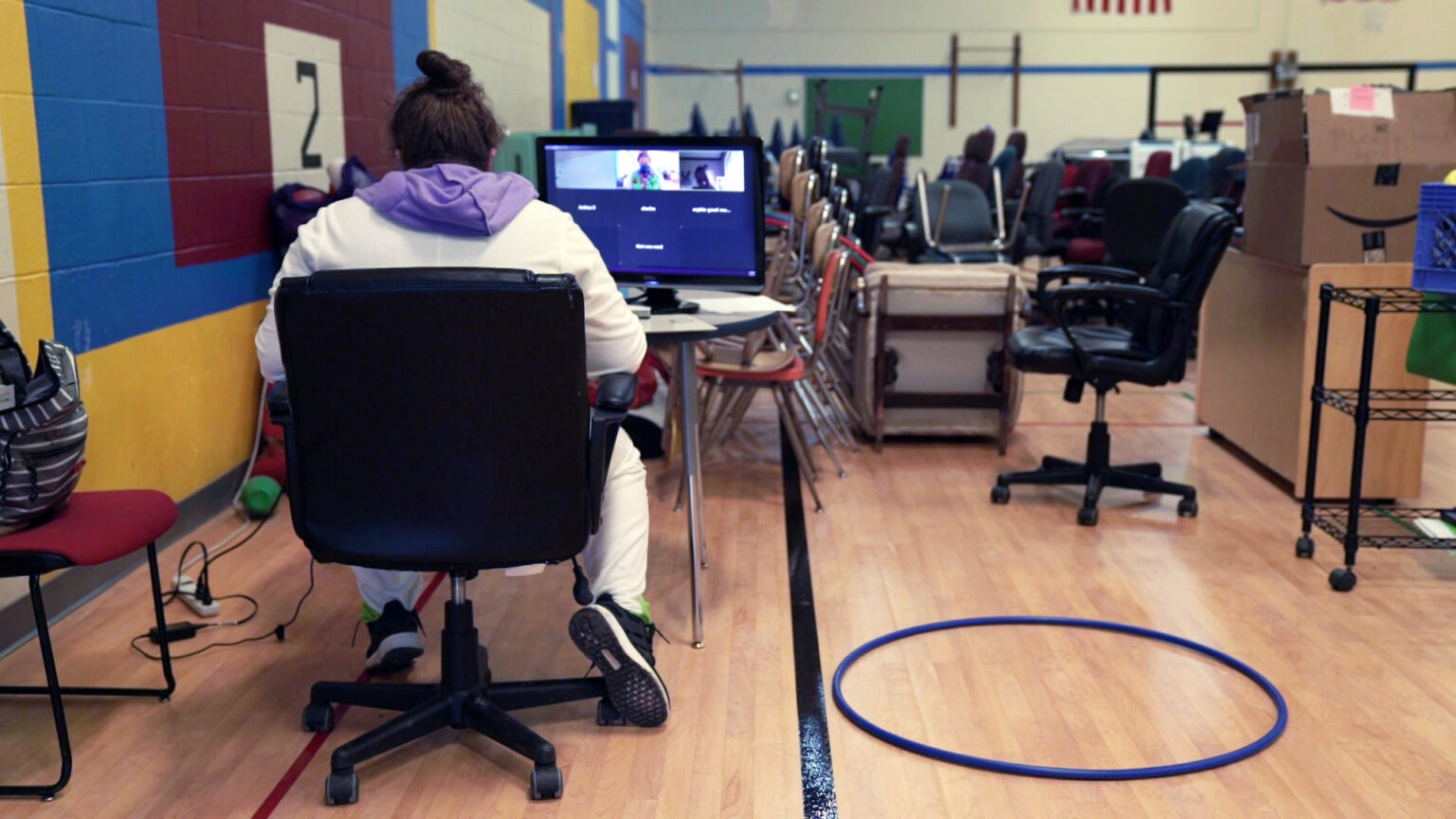 A teachers sits in an officer chair facing a screen for a virtual classroom in a gym filled with stacked chairs and other items.
