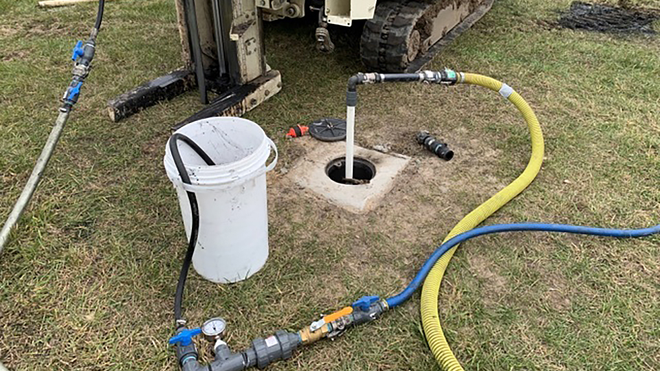 A treaded vehicle, a bucket and multiple pipes are connected on grass turf and connected to a drill hole.