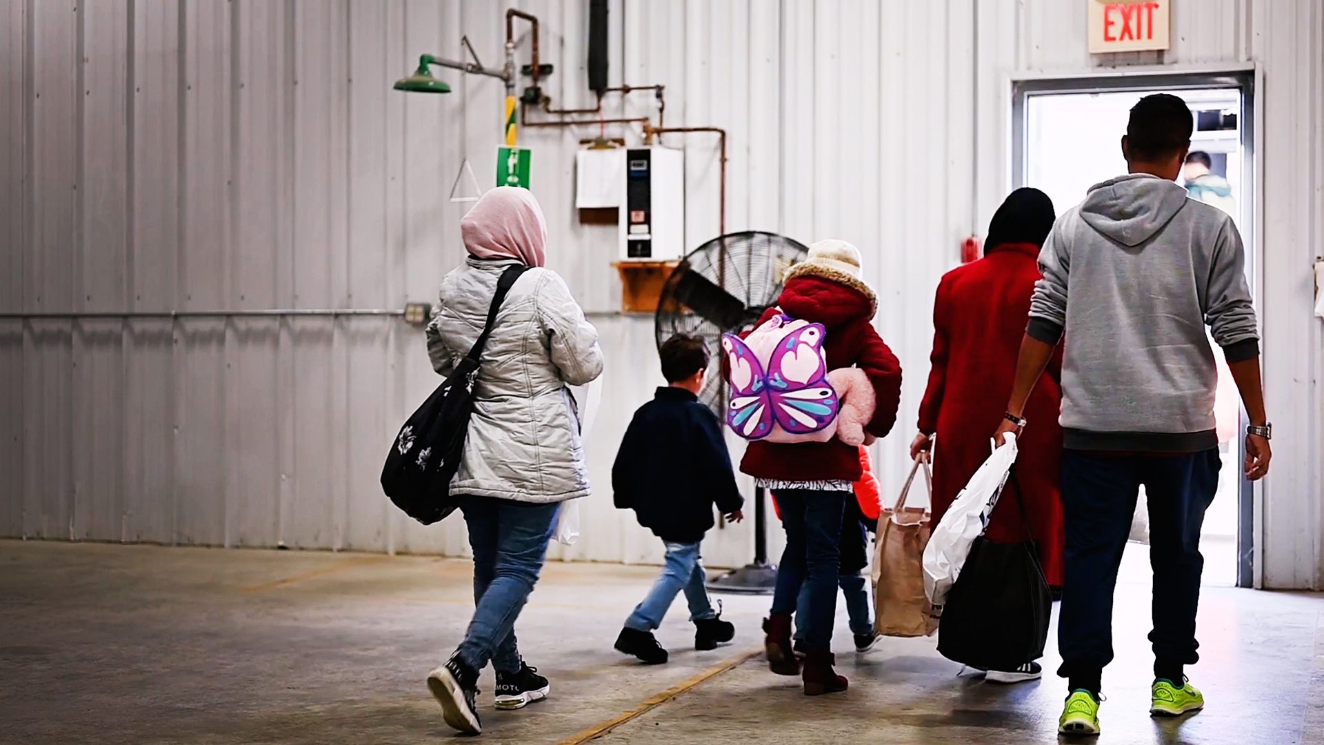 Multiple adults and children walk toward an exit door while inside a steel building with a concrete floor.