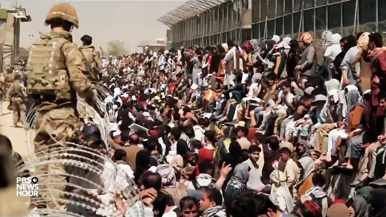U.S. troops guard hundreds of Afghans seated and standing behind razor wire.