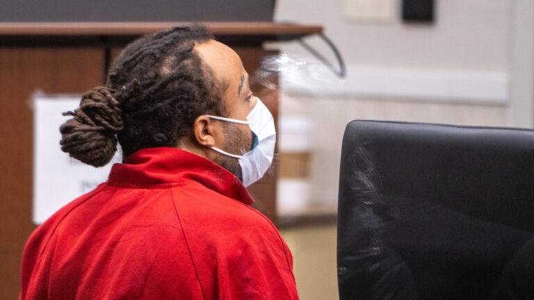 Darrell E. Brooks, seen through as glass window pane, sits in a courtroom while wearing a medical mask.