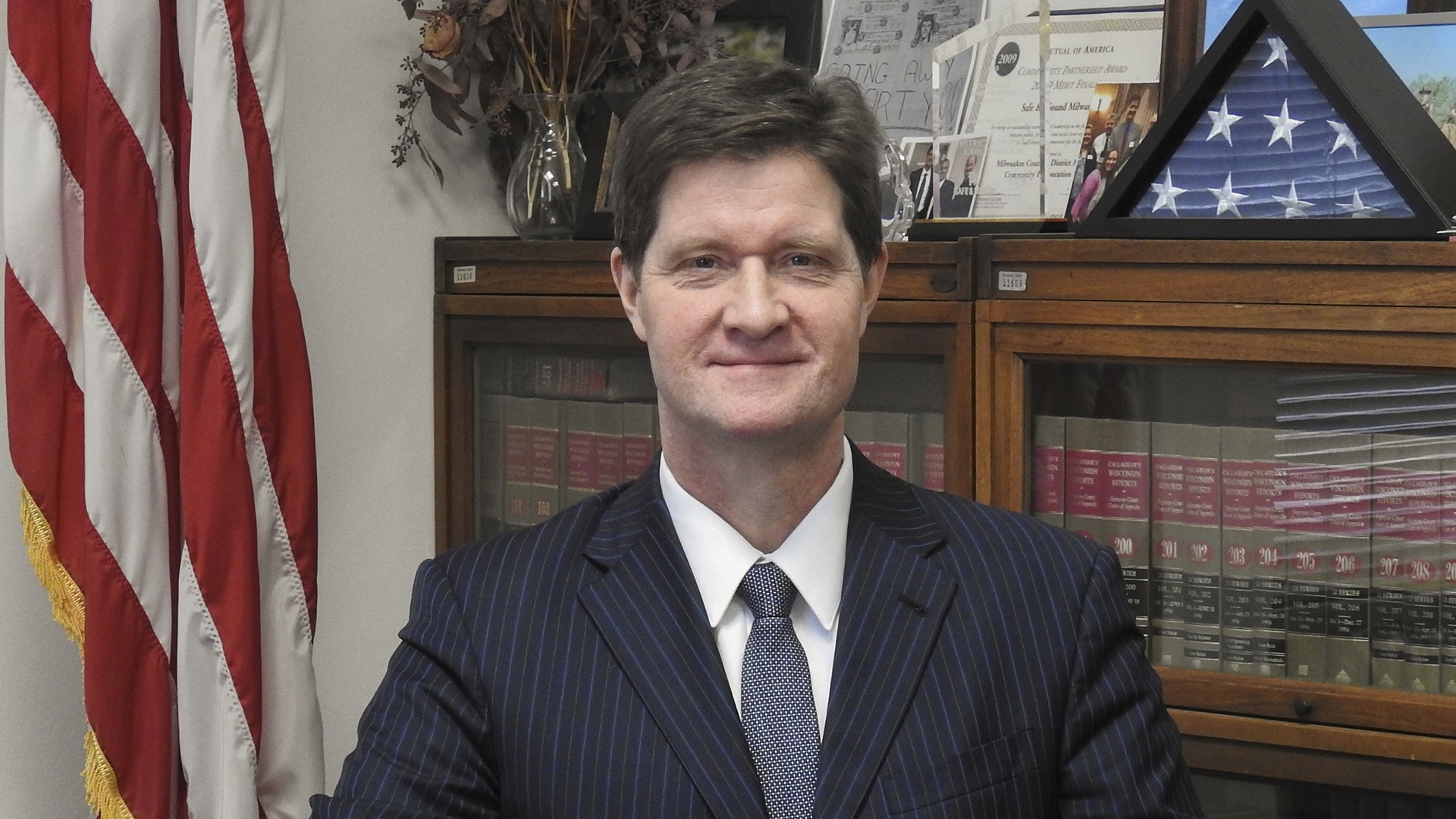 John Chisholm sits in an office with legal books in a cabinet, flags and other items in the background.