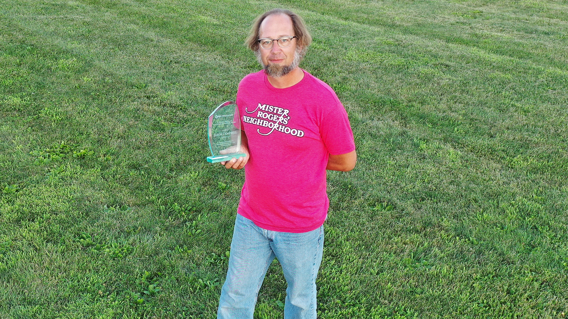 Jason Campbell stands in a field wearing a Mister Rogers' Neighborhood red t-shirt and jeans, holding his 2020 Volunteer of the Year trophy/award