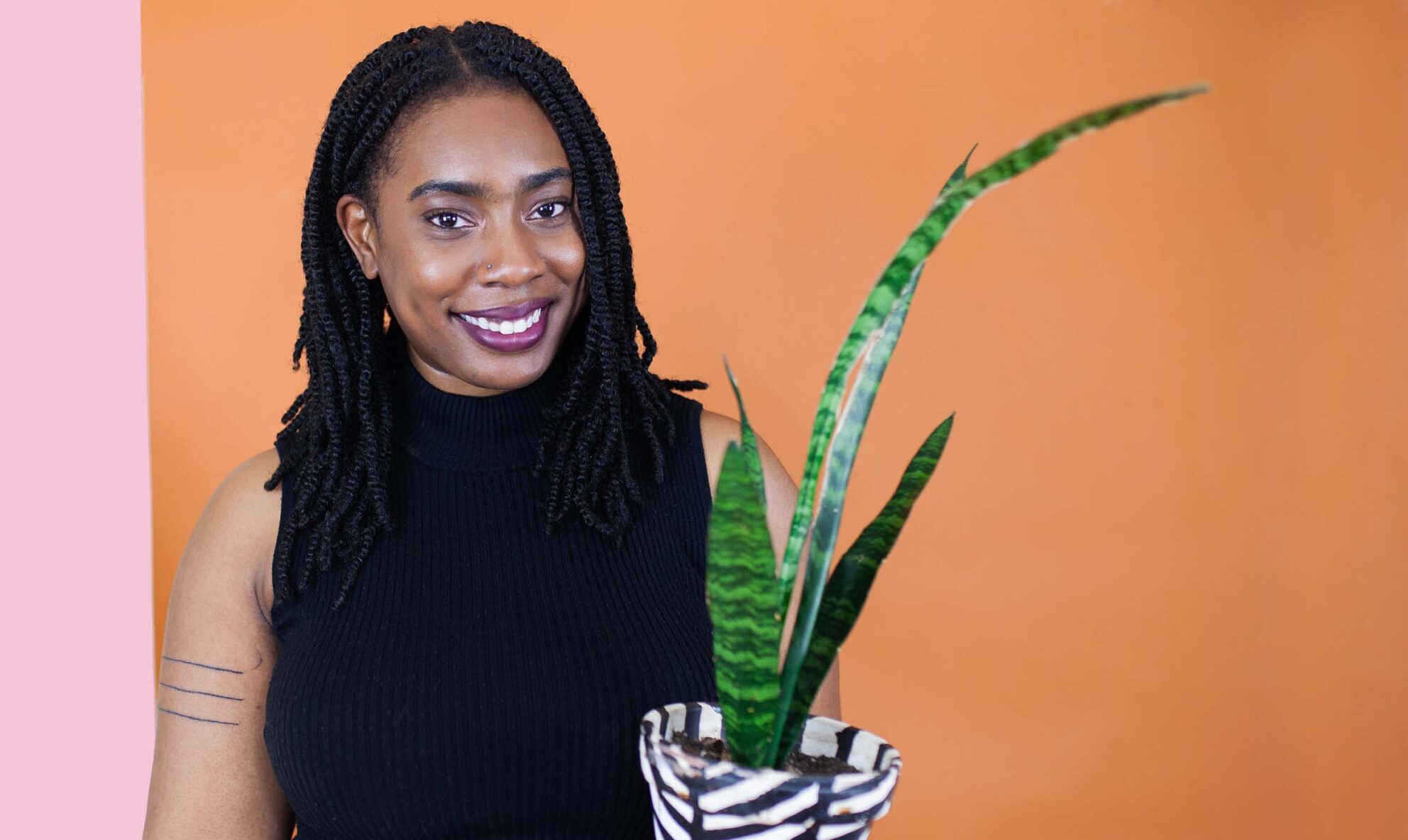 Quantese Winters stands in front of a pink and orange wall smiling and holding a houseplant