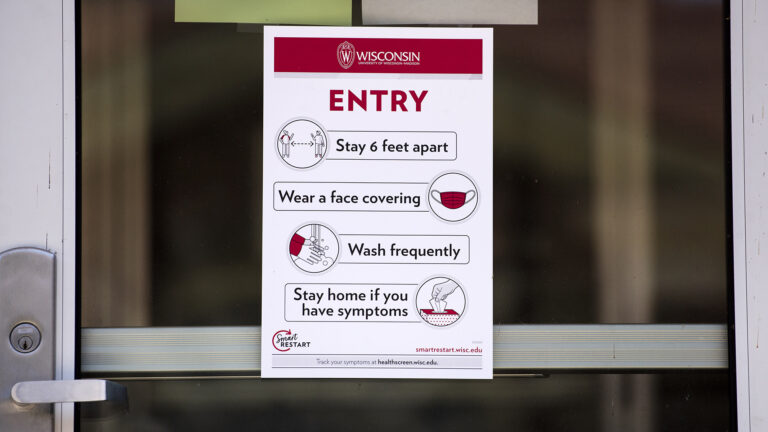 A paper sign attached to a glass door with the UW-Madison seal reads ENTRY with specific precautions of Stay 6 feet apart, Wear a face covering, Wash frequently, and Stay home if you have symptoms with accompanying graphics.