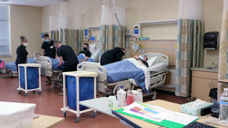 An instructor teaches Wisconsin National Guard troops wearing medical scrubs how to care for patients in a simulated hospital setting with beds and other medical equipment.