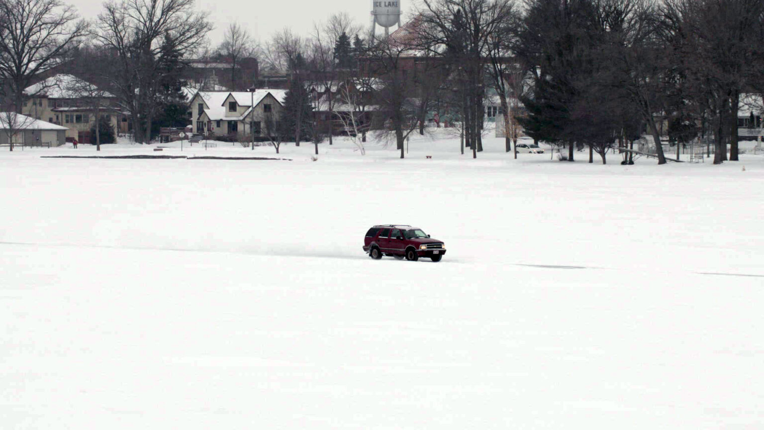 An SUV drives on an ice road on a lake with trees and buildings on the shore in the background.