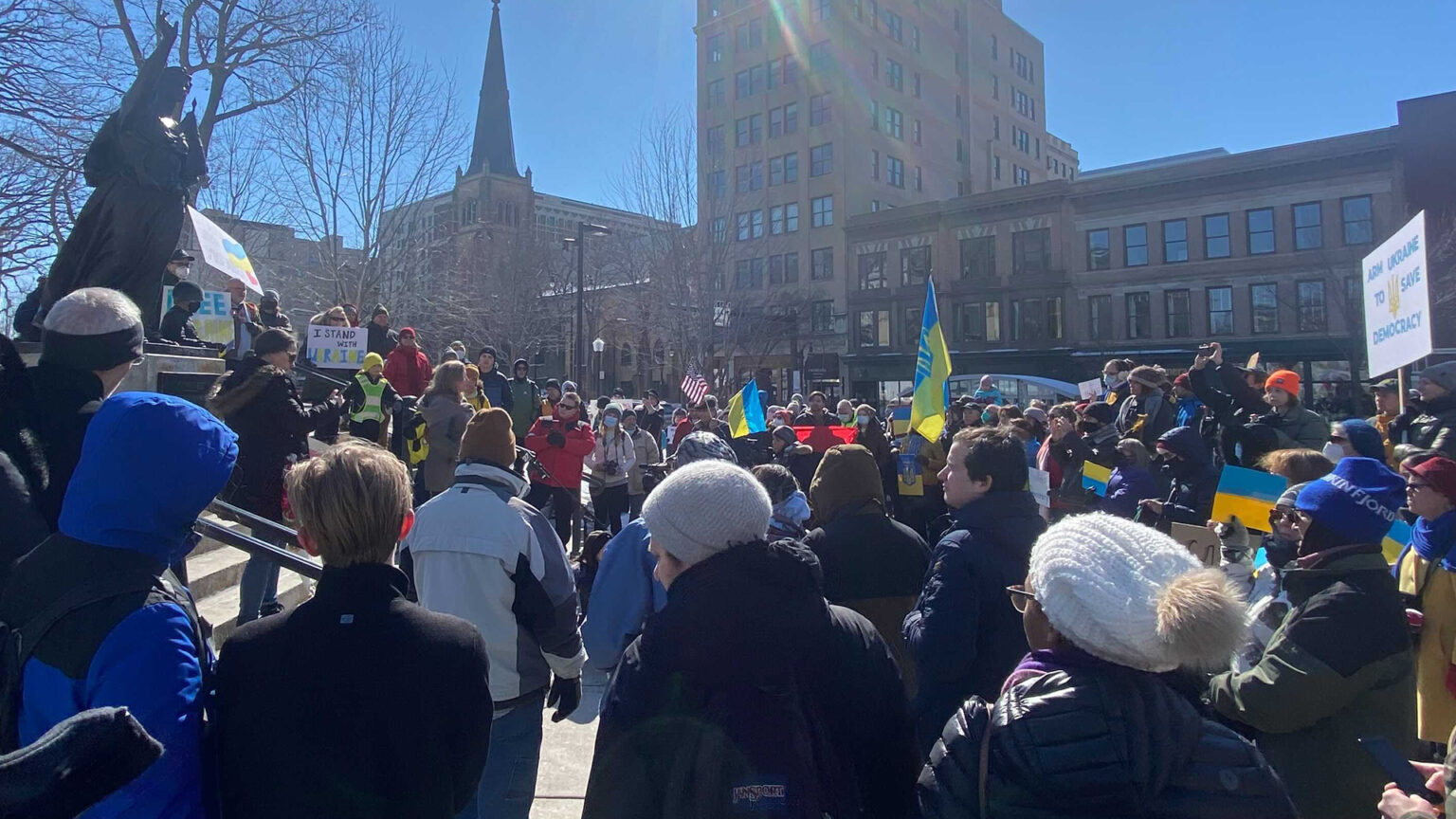 A crowd gathers below a statue holding Ukrainian and American flags and signs with messages supporting Ukrainians.