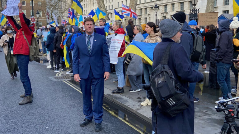 Ron Kind stands on a street with demonstrators holding signs and the flags of Ukraine and the United Kingdom on the sidewalk in the background.