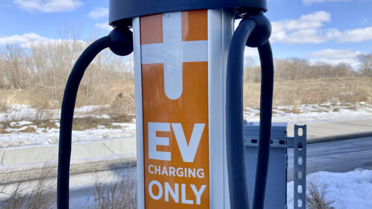 An electric car charging station with connection cords stands in front of a road, sidewalk and field with some snow on the ground.