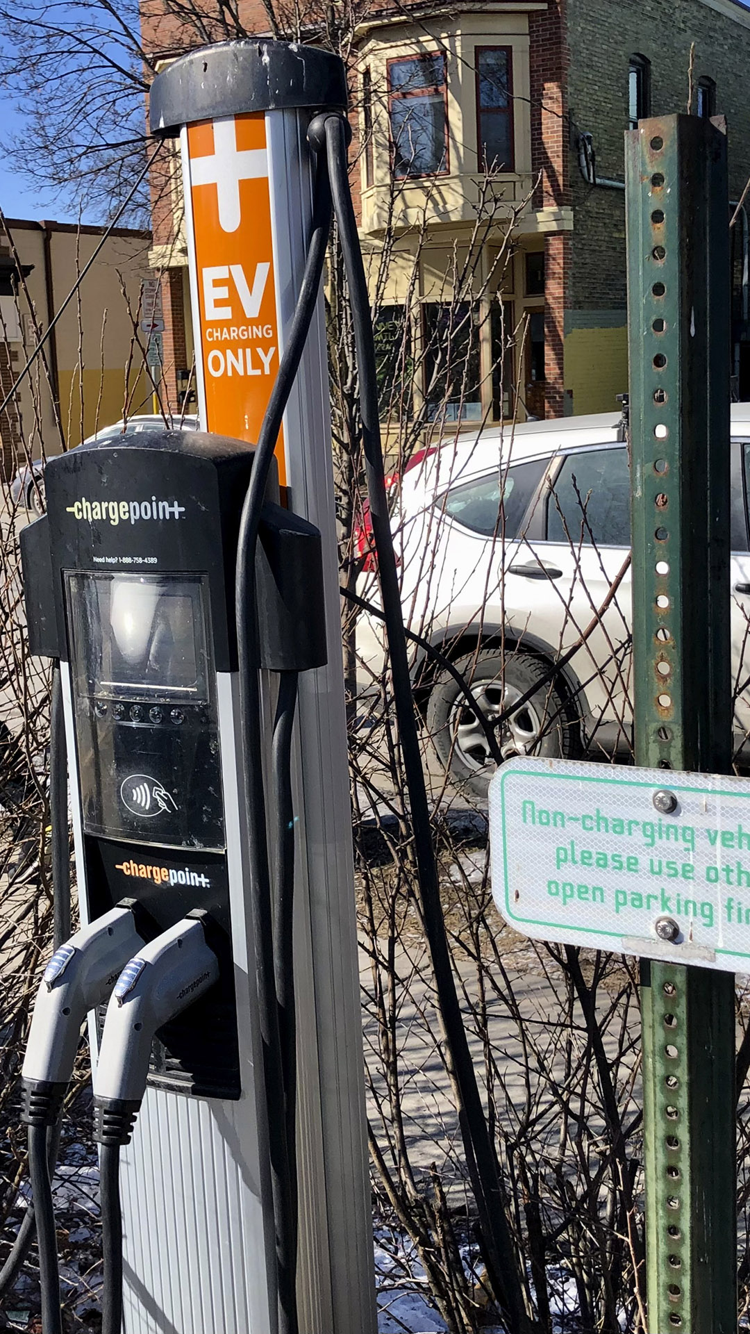 An electric car charging station with connection cords stands in front of a road, sidewalk and surrounding buildings.