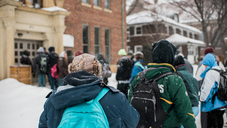 People wearing winter clothes and backpacks stand in line in the snow outside a brick building.