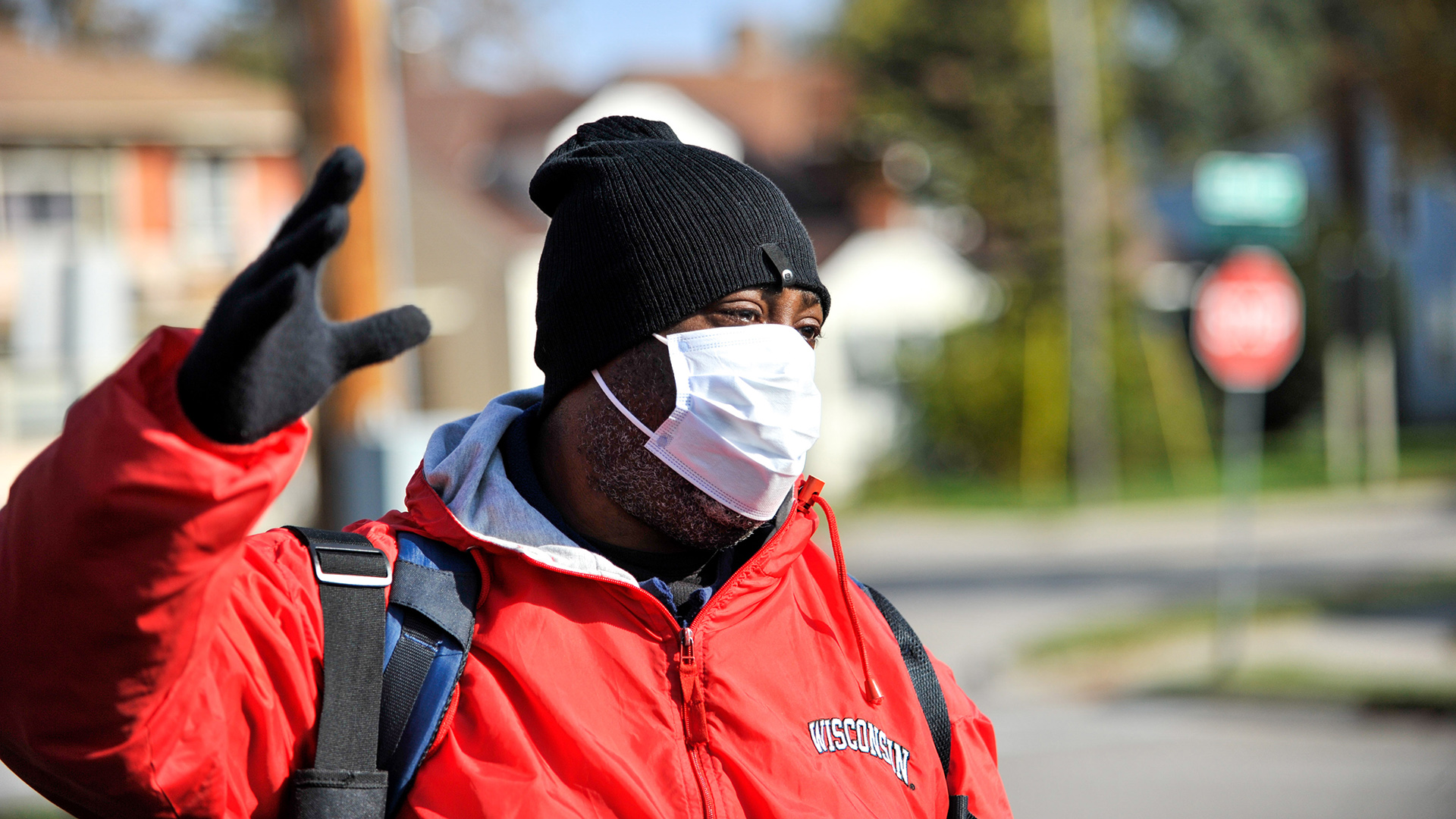 Madee wears a Wisconsin Badgers jacket, face mask and stocking cap, standing next to a street and holding a hand up in the air.
