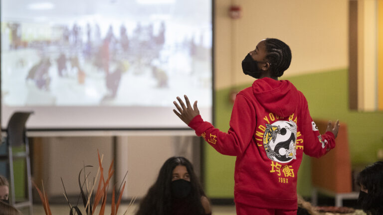 Terrion Gonzalez stands with outstretched arms inside a classroom, wearing a mask and with other people seated nearby.