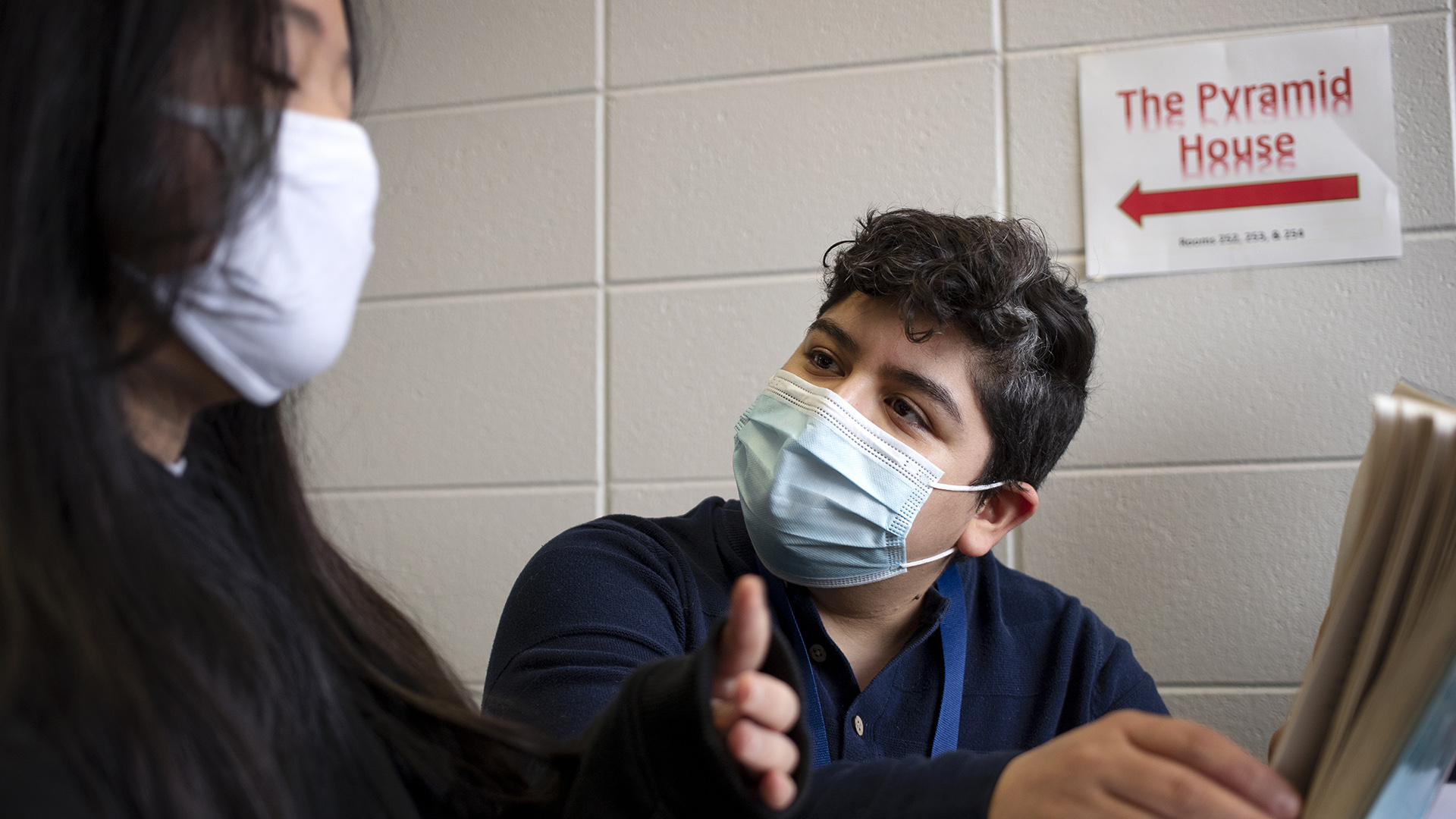 Steph Guzman and a student, both wearing pandemic masks, speak to each other about a book.
