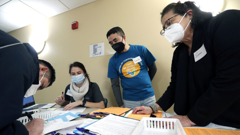 Claire Holzschuh, Long Vue and Susana Guerra sit and stand on one side of a table topped with pens and papers, helping another person fill out a form.