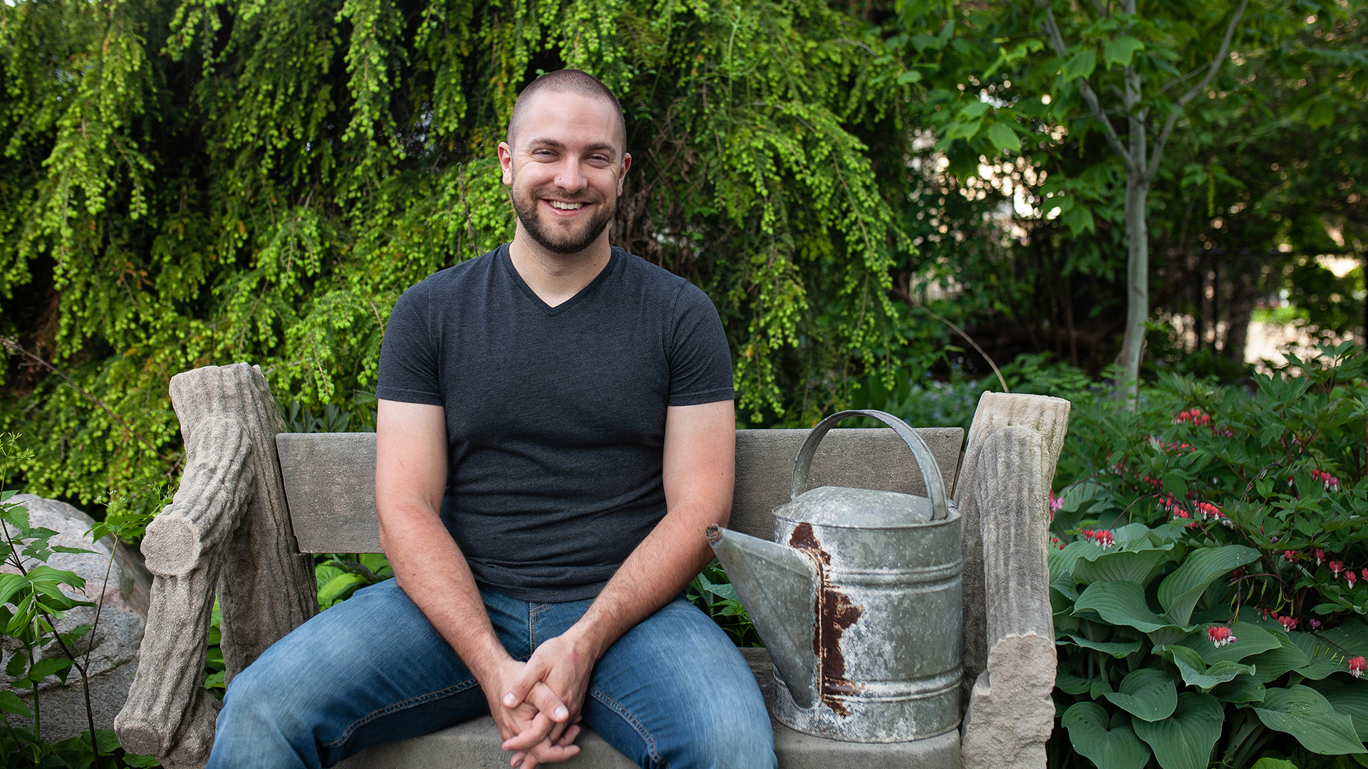 Co-host Ben Futa sits on a garden bench next to a weathered watering can with green shrubs and trees behind and around him
