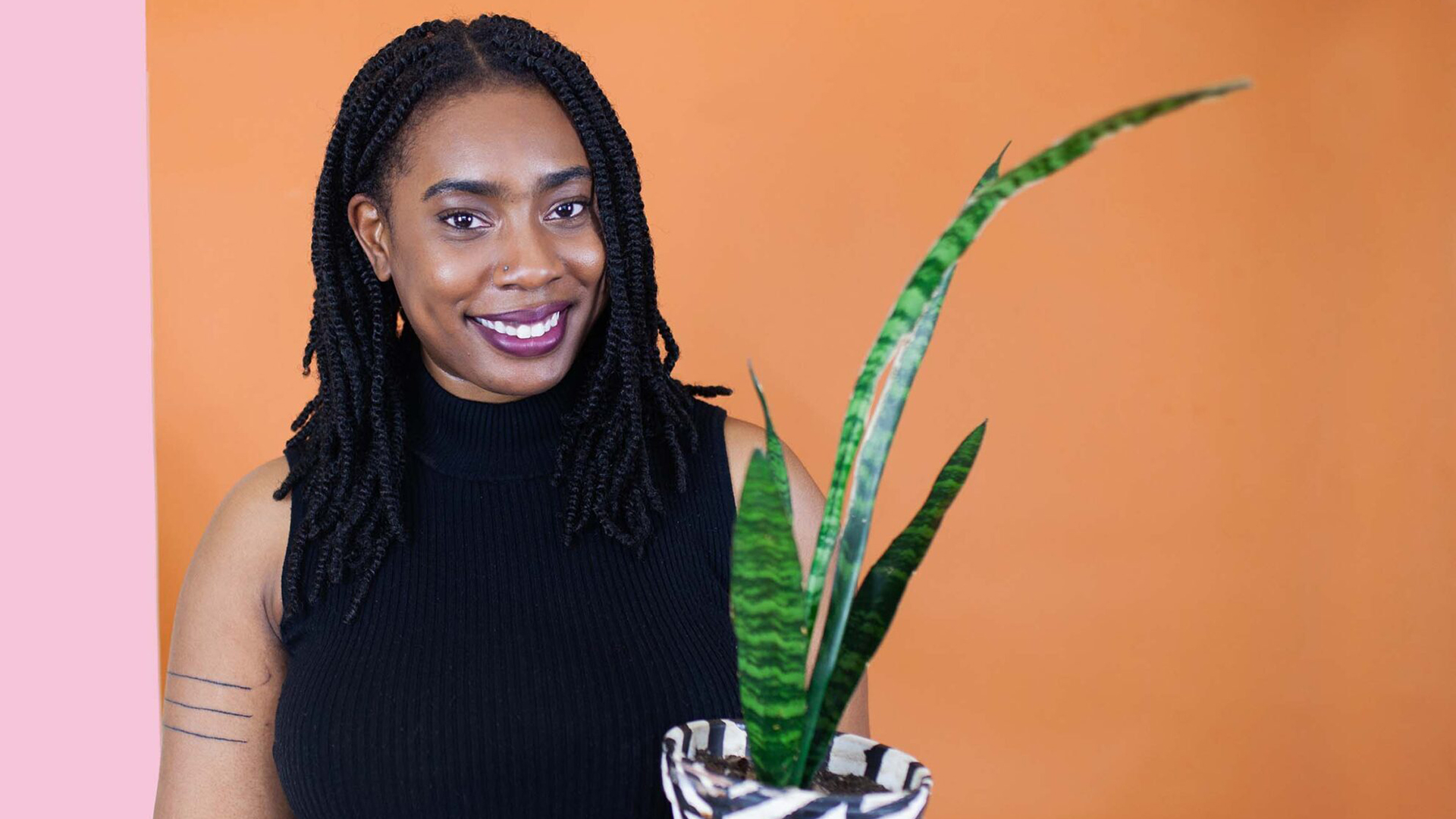 Co-host Qwantese Winters holds a house plant while standing in front of a vibrant pink and orange wall