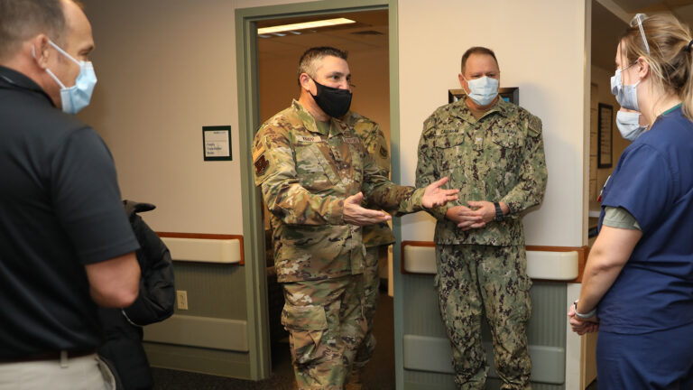 Paul Knapp, wearing a field uniform and a face mask, speaks with other individuals in field uniforms and medical scrubs in a hospital hallway. 