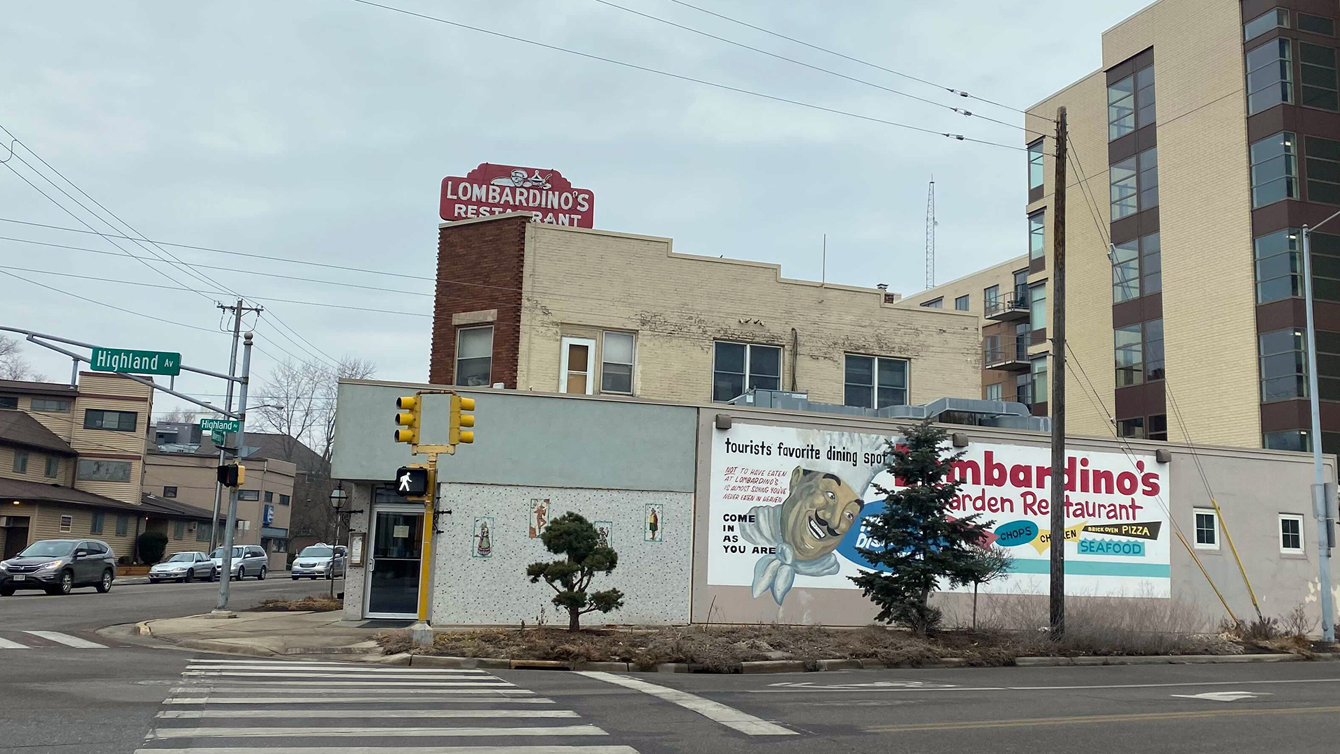 A mural advertising "Lombardino's Garden Restaurant" is painted on the side of a low-slung building, with taller buildings standing in the background and cars driving on the adjacent street.