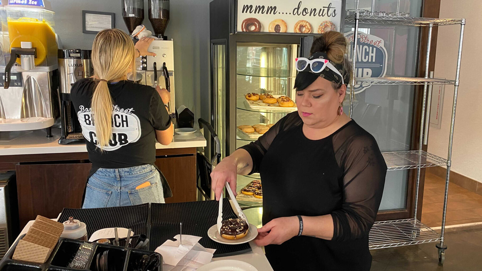 Dina Martinez uses tongs to place a donut on a small plate while another restaurant worker prepares a coffee drink.